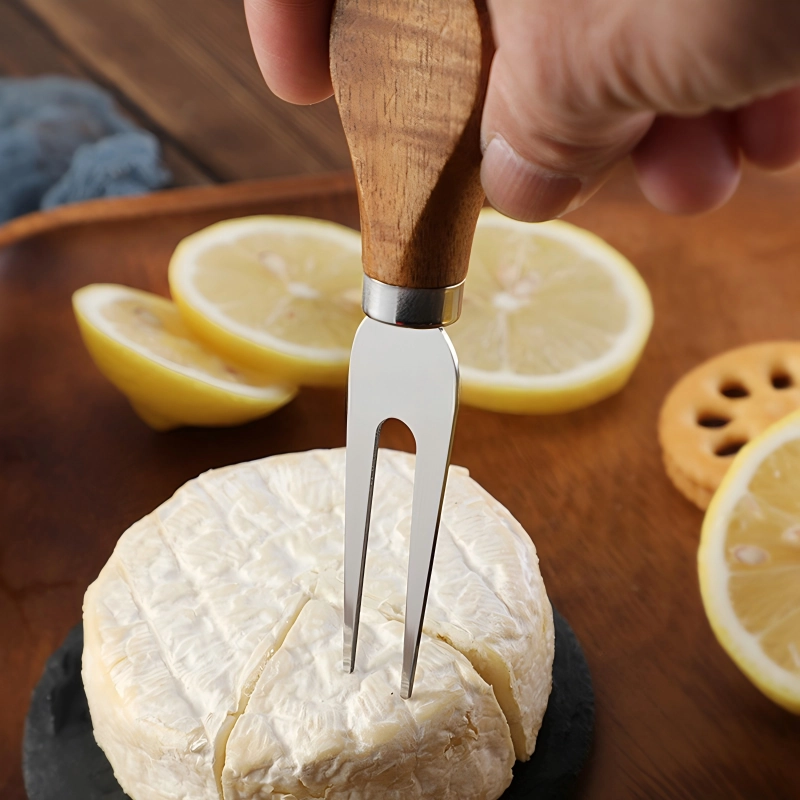 2-tine stainless steel cheese fork with wood handle piercing brie cheese displayed on wooden tabletop, KL-KA-GAD-007.