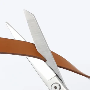 All-metal stainless steel tailor scissors cutting through a brown leather strap isolated on white background, SKU: KL-SC-HC-001.